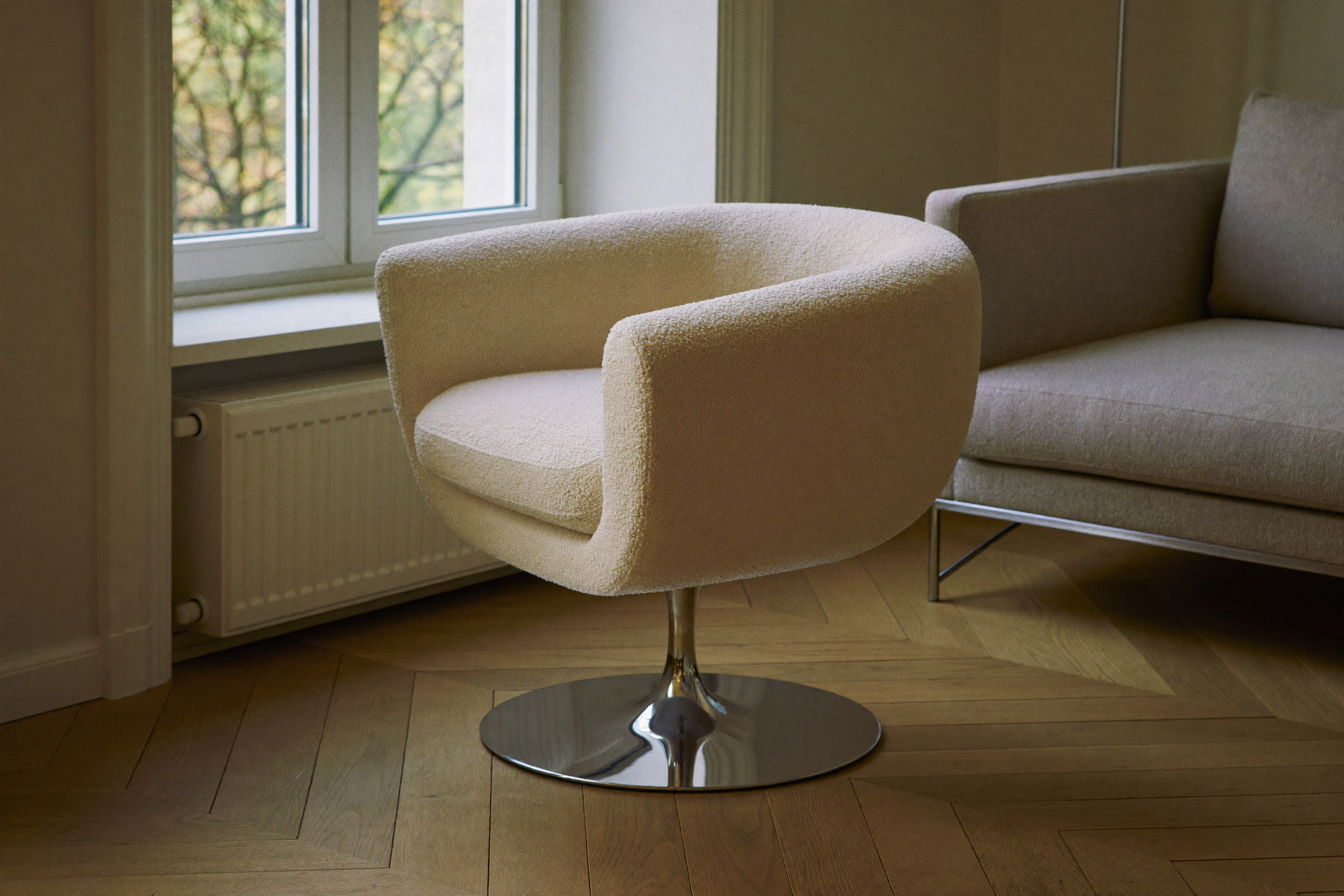 Beczkowy fotel obrotowy w kremowym bouclé na chromowanej podstawie talerzowej, salon z naturalnym światłem i podłogą z desek