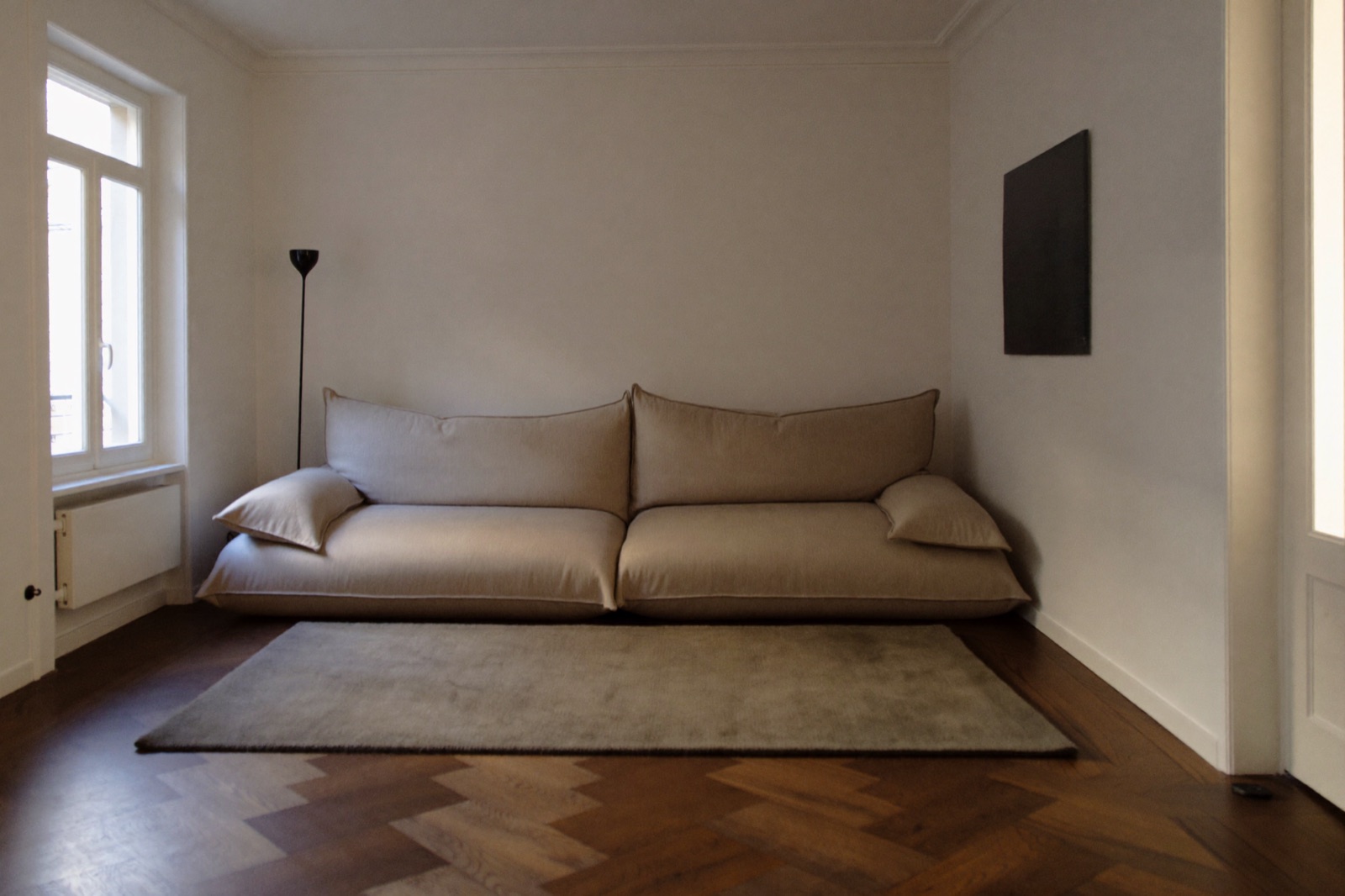 Duża beżowa sofa zajmująca całą ścianę niewielkiego pokoju, obok jeden duży dywan i wysoka lampa podłogowa